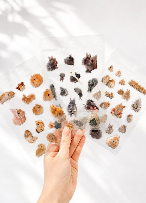 A hand holds three clear plastic sheets filled with small animal-shaped stickers or charms, including rabbits, squirrels, and other furry creatures, against a bright white background with soft leaf shadows.
